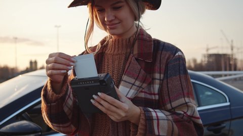Cowboy girl in country outfit smiling while taking selfie pictures on polaroid. Joyful young woman checking fresh retro instant photograph near parked car at sunset.