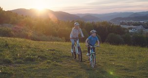 Mother and son ride a bike. Happy cute boy in helmet learn to riding a bike in park on green meadow in summer day at sunset time. Family weekend. 4K video, Slow motion
