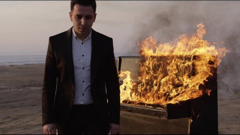 A serious man in a classic suit and white shirt amid a bright flame-burning piano on a sandy beach at sunset goes away, looks into the camera. art scene, art production