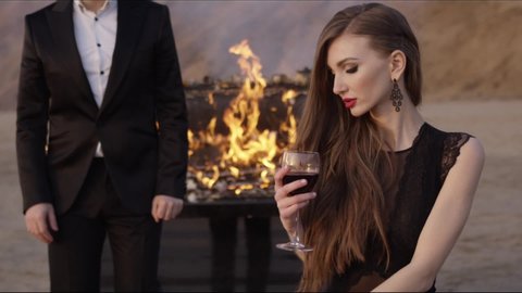 A young beautiful woman with a make-up and long hair on a sand quarry sits with a glass of red wine, a man in a classic black suit passes by. In the background is a flaming piano