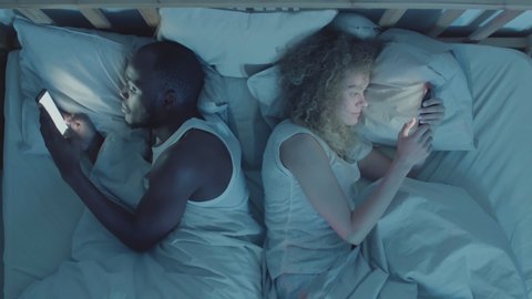Directly above view of African man and mixed-race woman lying back to back in bed with telephones in their hands