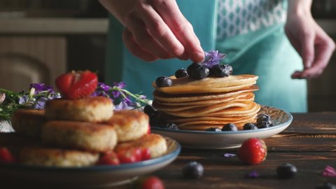 Female chief puts dish with fresh cocked blueberries and strawberries pancakes for breakfast in the morning light on a dark wooden table. Healthy Breakfast concept. Selective focus.