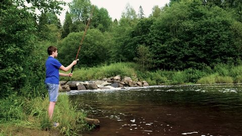 teenager boy stands on the riverbank with a long fishing rod. fishing. side view. water threshold, waterfall. summer.
