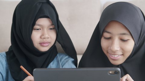 Two Muslim girl is studying online via the internet on tablet in living room at home, Asian elementary school children watching computer tablet. Concept of education at home. Close-up on the face kid