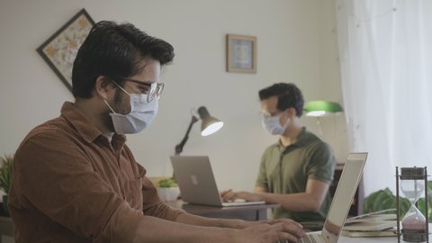 Two male or men office or corporate employees taking precautionary safety measures by wearing a protective face mask and maintaining social distancing while working on laptops in an indoor set up
