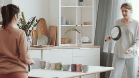Two young cheerful women playing using frying pans as racquets and tea mugs instead of net while playing ping pong at kitchen table while staying at home under quarantine - Powered by Shutterstock - Get 15% off with code: PIKWIZARD15