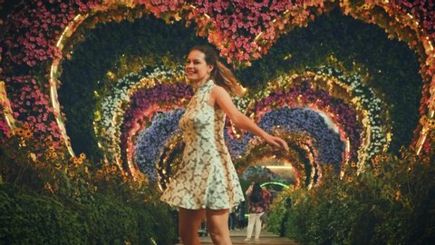 Happy woman tourist, walks along romantic evening alley in blooming Garden of Dubai city. Whirls dancing. smiling face. Landscaping design flowers arch in shape of hearts. night garland illumination
