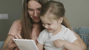 Mother with little cute daughter having fun and laughing using tablet. Funny games app. Mum with child take selfie, watch cartoons, make video call sitting on sofa at home.