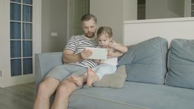 Father with little cute daughter having fun and laughing using tablet. Funny games app. Dad with child take selfie, watch cartoons, make video call sitting on sofa at home.