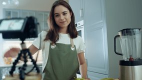 Attractive Caucasian Girl Recording Video Using Digital Camera on Cooking Healthy Food. Young Female Vlogger Filming in Kitchen at Home, Waving Hello and Talking to Her Audience. Slow Motion Shot