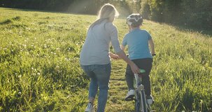 Mother teaching son to ride bicycle. Happy cute boy in helmet learn to riding a bike in park on green meadow in summer day at sunset time. Family weekend. 4K video