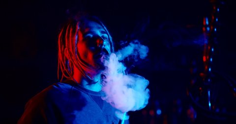 Hipster guy with dreadlocks looking at camera while smoking a hookah in neaon light, exhaling clouds of smoke - nightlife, tobacco concept 4k footage close up portrait