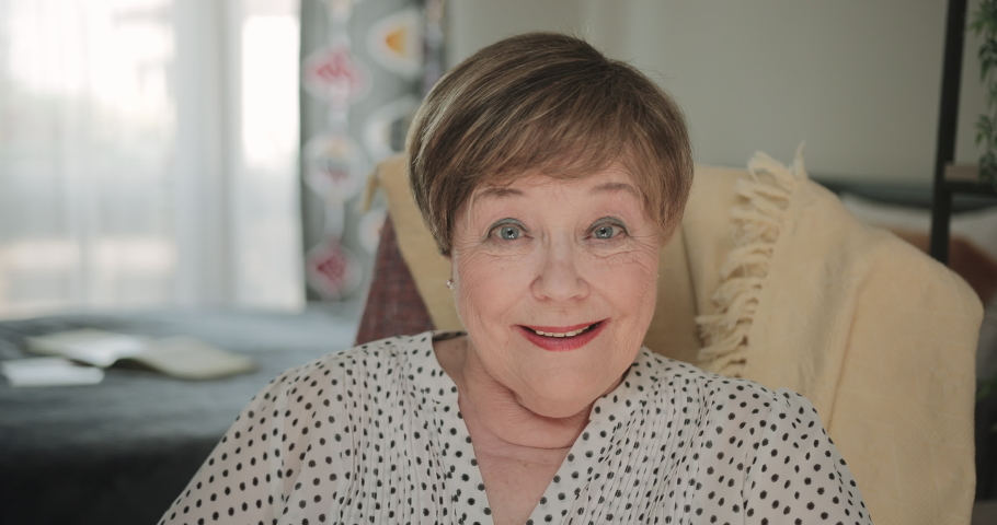 Close up view of mature woman pensioner sitting on chair at her apartment. Portrait of beautiful elderly lady smiling while looking to camera. Concept of peole and positive emotions
