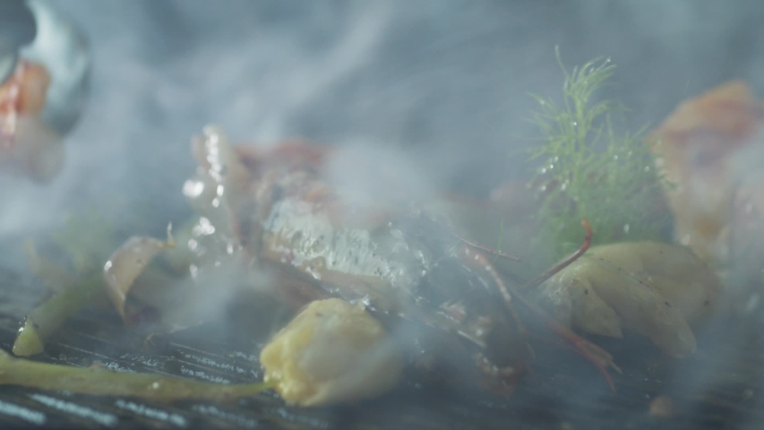 Сhef with tongs flips the prawns that are grilled with herbs and garlic in thick smoke with olive oil close-up Royalty-Free Stock Footage #1053189938