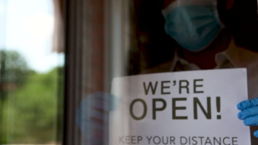 Business owner with gloves and face mask hanging an open sign  "We're open. Keep your distance" at a restaurant. Concept of new normality due to coronavirus | Shutterstock HD Video #1053190940