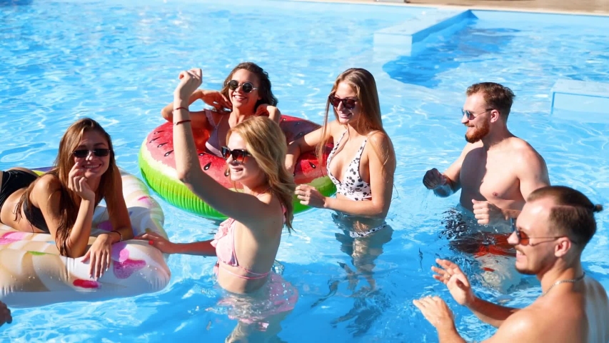 Friends have party in a private villa swimming pool. Happy young people in swimwear dancing, bonding and clubbing with floaties and inflatable mattress in luxury resort on sunny day. Slow motion. | Shutterstock HD Video #1053193031