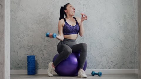Junk food. A young woman in sportswear is exercising with dumbbells and eating a high-calorie donut. The concept of diet and weight loss.