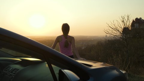 Young woman standing near her car enjoying warm sunset view. Girl traveler leaning on vehicle hood looking at evening horizon.