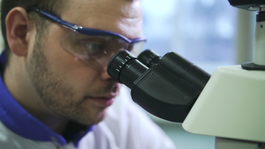 Male scientist is using microscope in working process in pharmaceutical laboratory spbd. Face view of young man researcher is looking through eyepiece of magnifying equipment and doing research in | Shutterstock HD Video #1053197930
