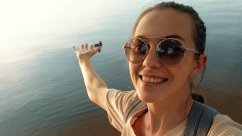 High angle joyful adult female in sunglasses and casual clothes smiling at camera while standing with reach out hand on lake beach against blurred wild duck floating on tranquil water in sunny evening