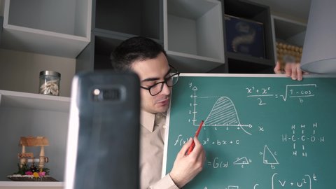 A male Caucasian online educator , holding a chalk board , teaching mathematics to his students via video call on  his smartphone mobile device