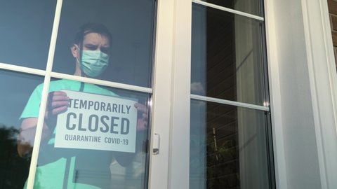 Temporary business closure. Caucasian white business owner wearing face mask attaching TEMPORARILY CLOSED sign at shop entrance due to coronavirus covid-19 epidemic outbreak over the world. 4K video