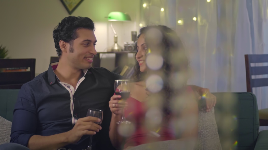 An attractive or beautiful couple sitting on a couch drinking a glass of wine together on a romantic evening. A good looking man and woman enjoying each others company on valentines day in house, home Royalty-Free Stock Footage #1053205946