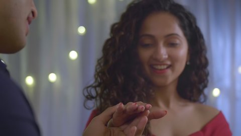 A young happy beautiful girl in red dress surprises and smiling when her boyfriend proposes by putting wedding ring in her finger. Excited female when male partner proposes for marriage. 