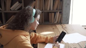Teen girl school college student wear headphones hold mobile phone distance learning online class math lesson with teacher tutor using zoom app video conference call write notes. Over shoulder view.