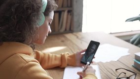 Teen girl school college student wear headphones hold mobile phone distance learning online class math lesson with teacher tutor using zoom app video conference call write notes. Over shoulder view. - Powered by Shutterstock - Get 15% off with code: PIKWIZARD15