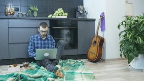 Male student sitting on floor at kitchen and working on laptop computer. Man works at home at remote workplace during pandemic. Man study online in internet, watching trainings, learning from home