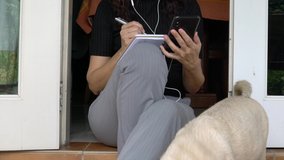 Woman writing on notepad and using cellphone. Home setting, with pug dog. Work from home concept.