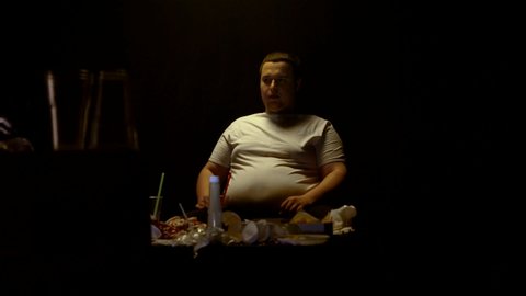 A fat man sits at home in front of the TV and looks at junk food. Gluttony and overweight, background