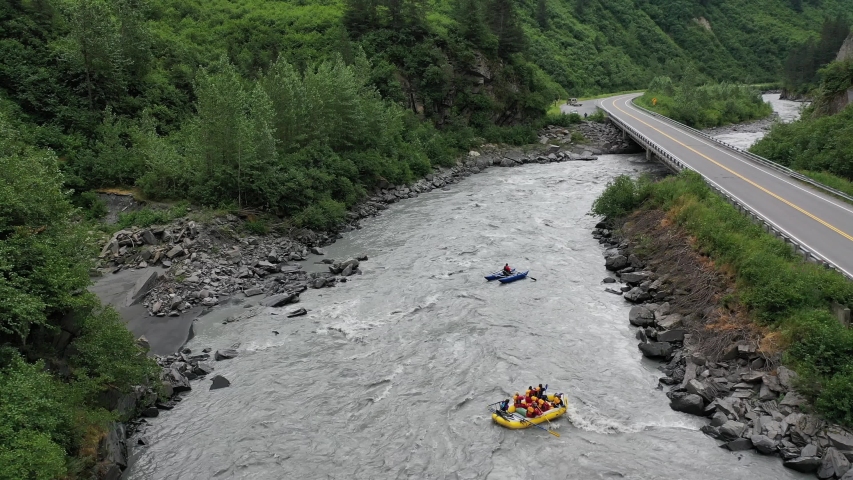 River raft sideways going down a raging river as they prepare to go under a bridge. Following Drone Shot with a safety boat in the back drop.