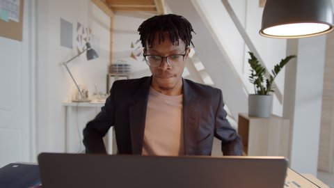 360-degree tracking shot of young African American businessman in smart casual outfit entering office, sitting at desk, opening his notepad and working on laptop