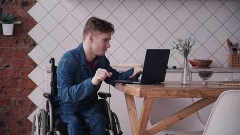 Man in wheelchair working with laptop at home. Adult disabled male working studying on laptop and checking news mail. sitting in modern kitchen interior. concept computer