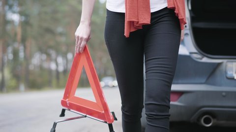 Unrecognizable Caucasian woman installing red triangle sign on suburban road. Misfortune on countryside road, female driver with broken automobile, car accident.