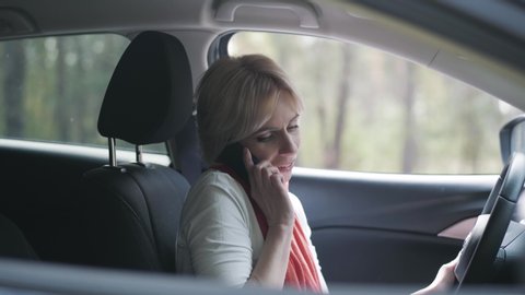 Portrait of confident female driver talking on the phone and applying lipstick. Side view close-up of beautiful blond Caucasian woman sitting on driver's seat. Lifestyle, positive people, happiness.