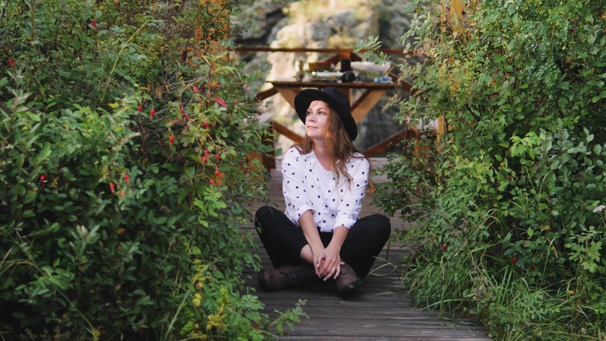 Attractive brunette in a hat and a white shirt with peas talking on a cell phone, taking a selfie on the phone, enjoying nature. A woman in a hat sits on the floor by the gazebo with a mobile phone.