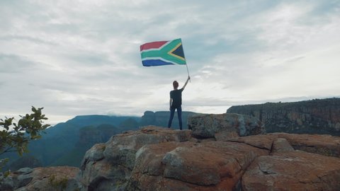 A slow motion shot of a young women standing on a cliff with the South African flag, overlooking a beautiful view.