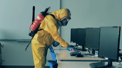 A man in a hazmat suit is disinfecting a computer class