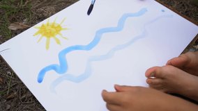 Closeup view video footage of little boy of 5 years old painting outdoors together with his mommy. Child paints blue sea water, sun, beach and sailboat on water on white sheet of paper.