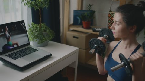 Young fit sporty brunette girl pumping muscles with dumbbells watching tutorial on laptop. Woman doing sports and fitness training at home during covid quarantine. Healthy lifestyle in pandemic time