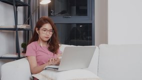 Young Asia teen girl student wears headphones distance learning lesson with online teacher on laptop in living room from home at night. Social distancing, quarantine for corona virus prevention.