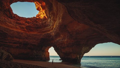 Cinemagraph - seamless video loop of a young model woman in a dress standing on the beach in the famous Benagil Cave at Algarve, Portugal at sunrise in summer with the waves of the sea moving gently.