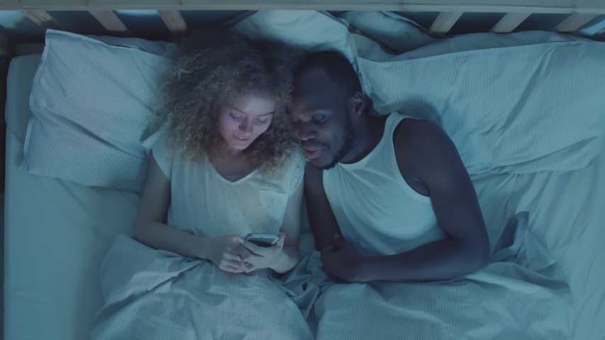 Directly above view of multiethnic young couple lying together in bed at nighttime, looking at cellphone screen and having discussion