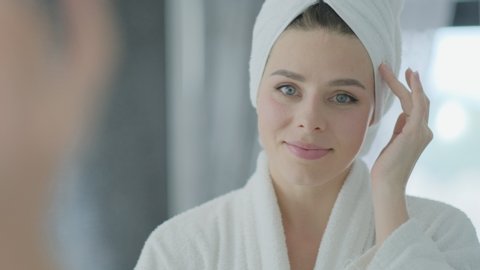 Close up portrait of young brunette woman looking in the mirror after shower at home facial skin care treatment, skincare concept.
