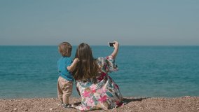 Back view of woman with a little boy making video call with parents and sitting on the shore by the sea on a sunny day. Mom and son spend their day together
