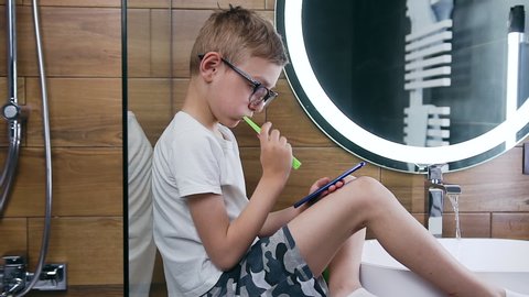 Portrait of good-looking blond 10-aged boy which wearing eyeglasses, dressed in homewear using his smartphone during brushing teeth with toothbrush in the bathroom