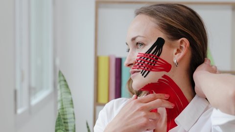 Beautiful woman with kinesio tapes on her neck and cheek against wrinkles looks at window and then to the camera. Facelift beauty procedure. Kinesiology treatment close up